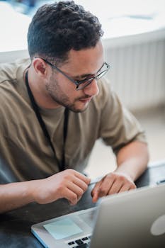 Adult man with eyeglasses and stubble working intently on a laptop in an indoor office setting.
