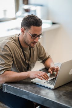 A man wearing eyeglasses works intently on his laptop in a modern office setting.