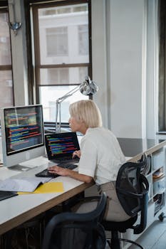 Blonde woman coding at desk with dual monitors in a contemporary office.