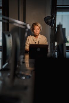 A woman working on a laptop at night in a dimly lit office. Warm lighting enhances focus.