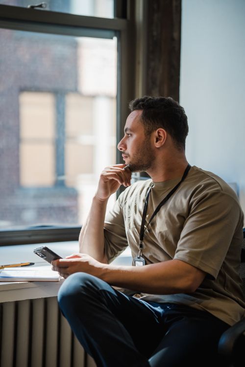 Free A man in an office sits cross-legged by a window, lost in thought while holding a mobile phone. Stock Photo