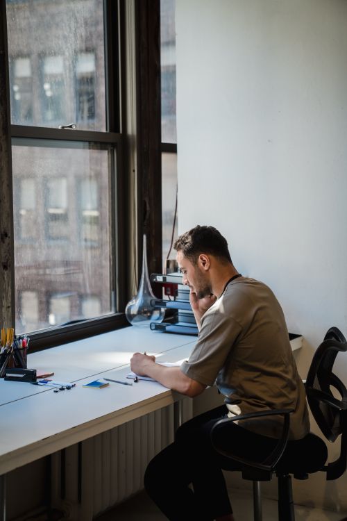 Free A man sits thoughtfully at an office desk by the window, engaging in phone conversation in a bright workspace. Stock Photo