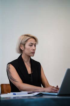 Blonde woman in office setting typing on laptop, showcasing tattoos and professionalism.