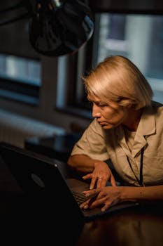 A woman concentrating on a laptop in a dimly lit office setting, showcasing dedication and focus.