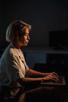 Blonde woman seriously focused on laptop, working late at night in dark office setting.
