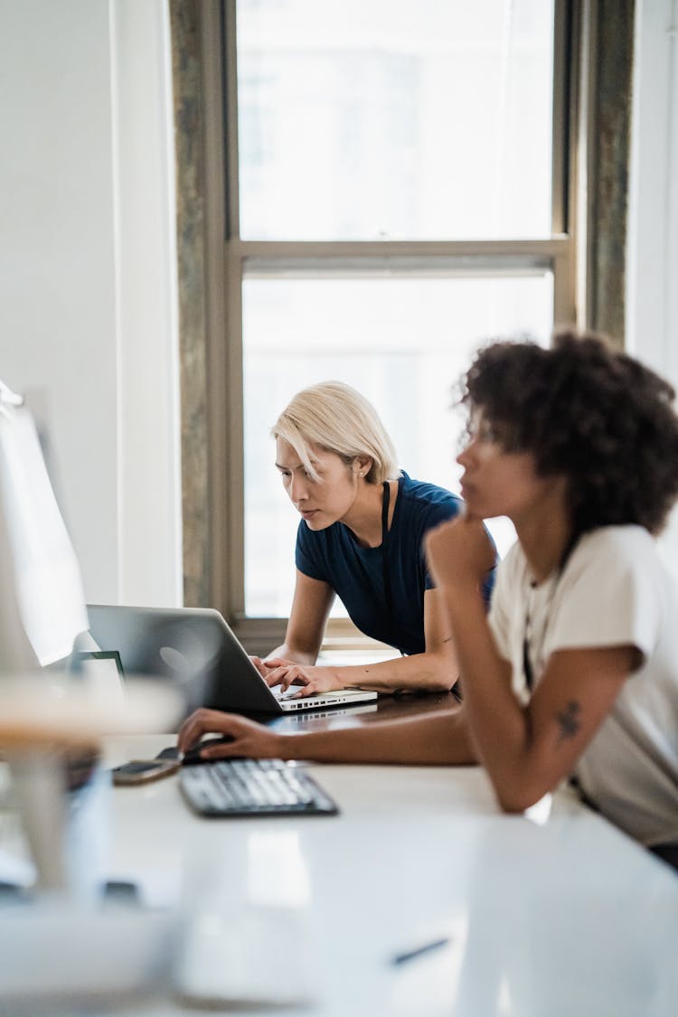 Two Women Sitting Next To Each Other At Office Desk And Working