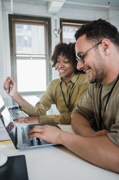 Two colleagues happily working on laptops together in a bright office setting.