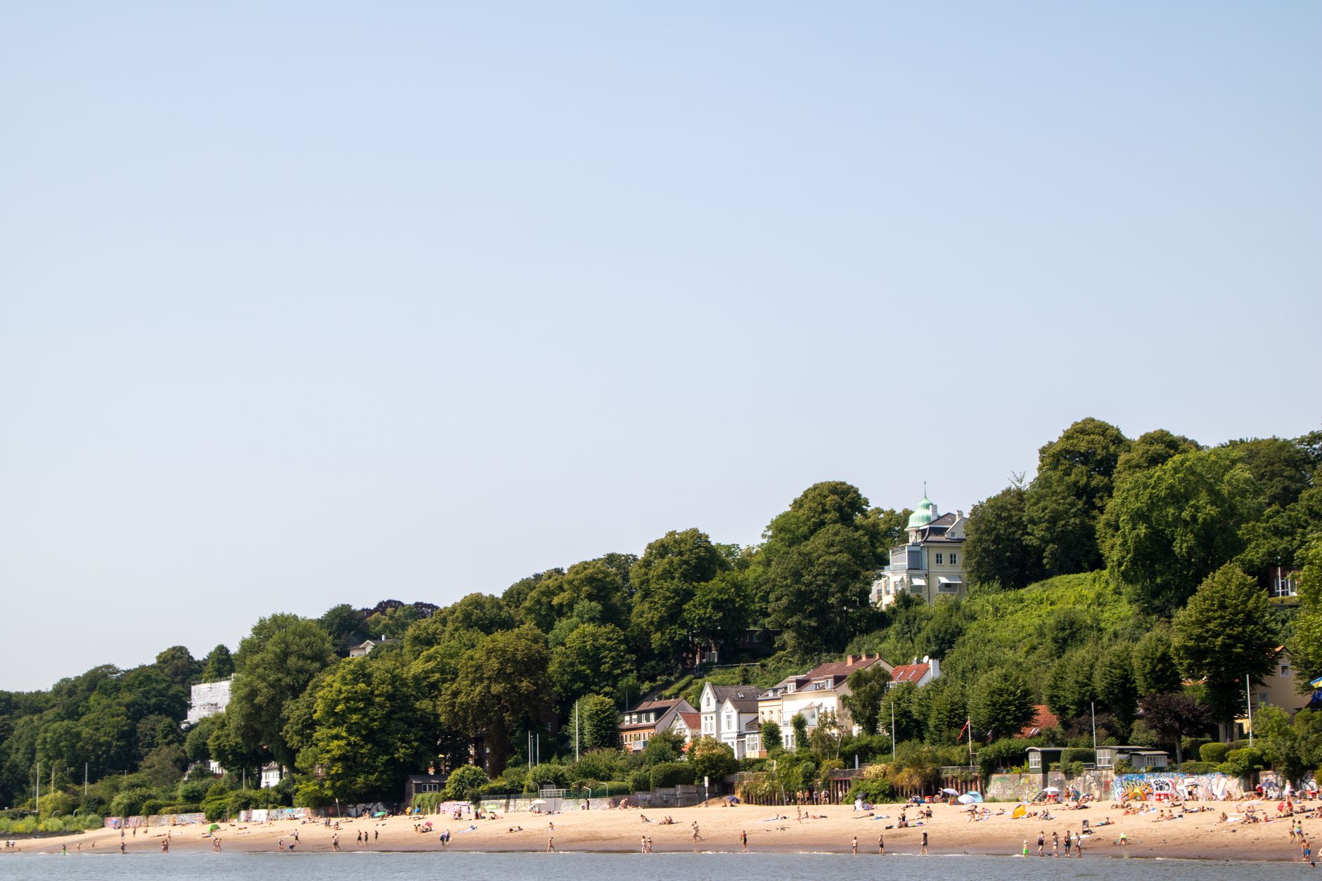 Sunlit beach with historic villas along Hamburg's coast, surrounded by lush greenery.