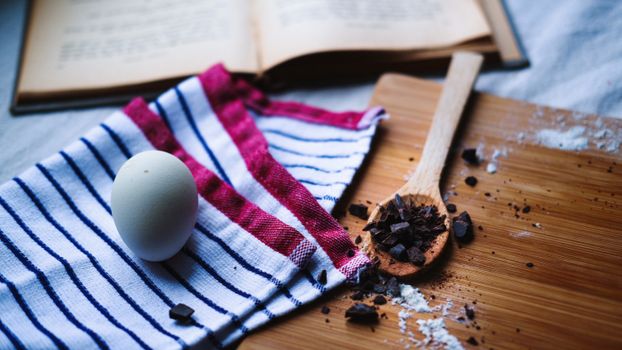 Overhead view of baking essentials with egg, chocolate, and open cookbook on wooden table.