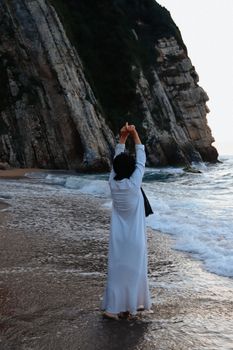A woman in a white dress standing on a rocky coastline near the water, facing the cliffs.