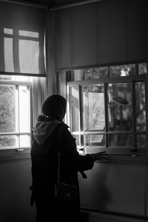 Grayscale Photo of Person Standing by the Window