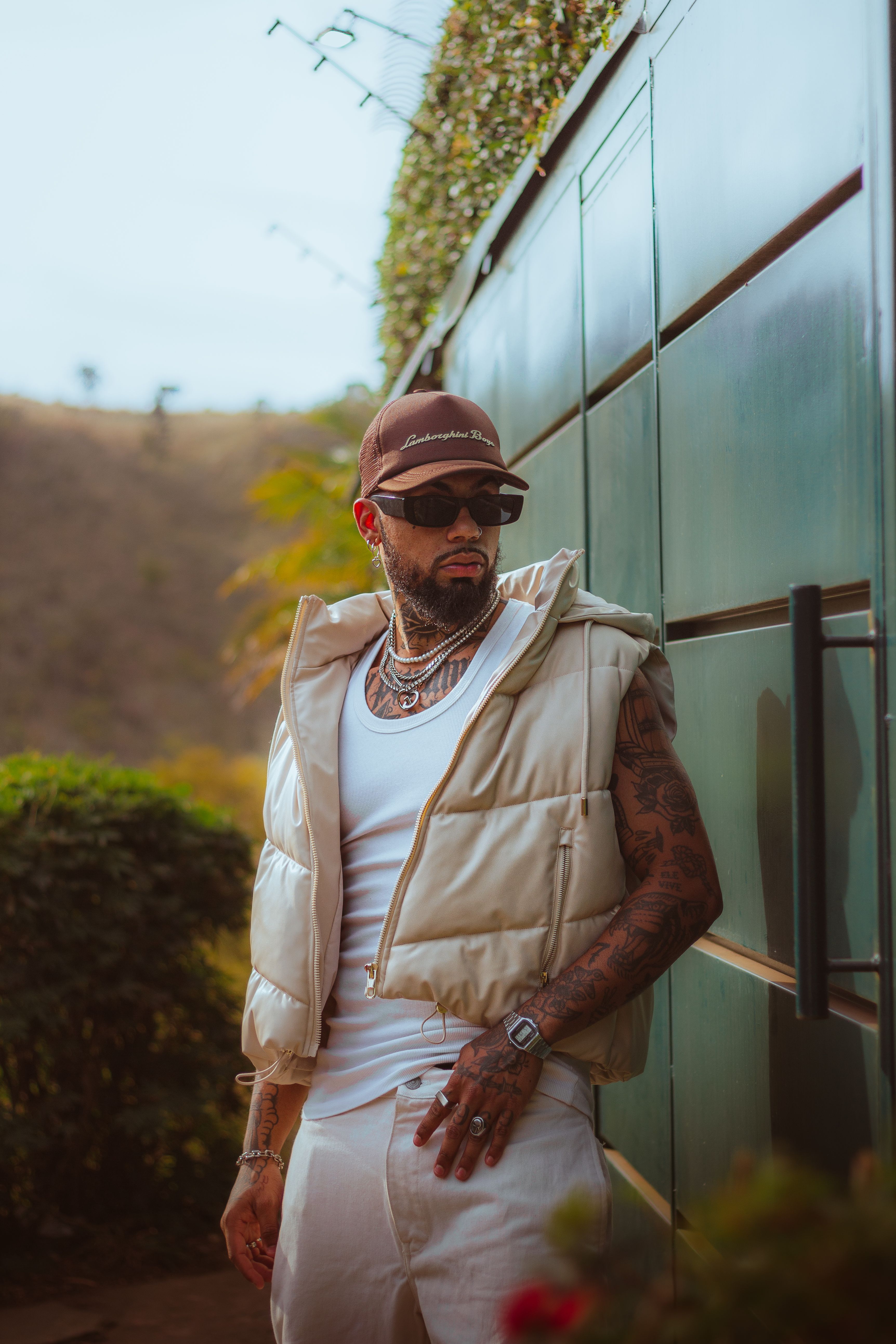Fashionable man with tattoos poses outdoors wearing sunglasses and a cap.