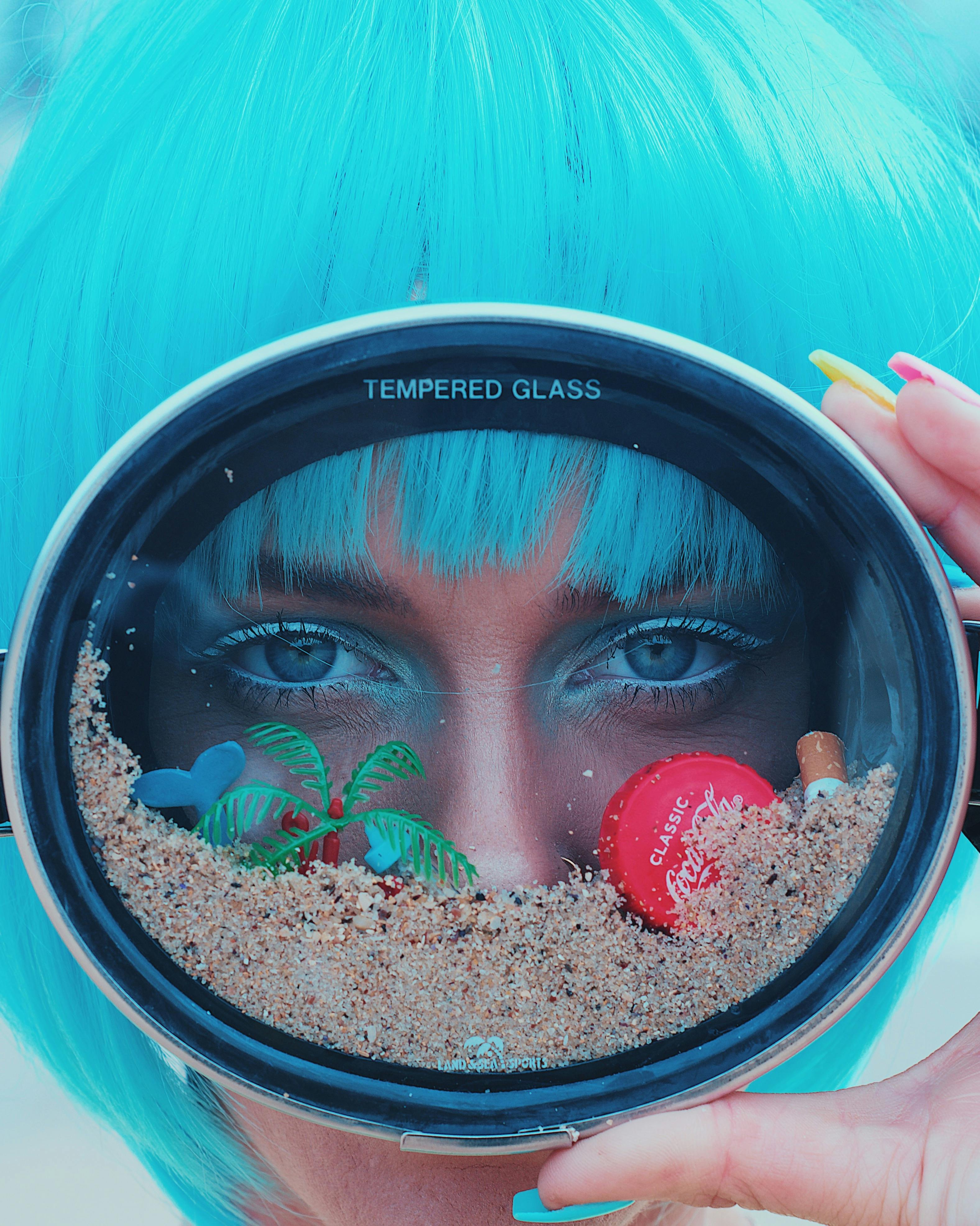 Artistic portrait of a woman with blue hair viewing through a tempered glass lens with sand and items.