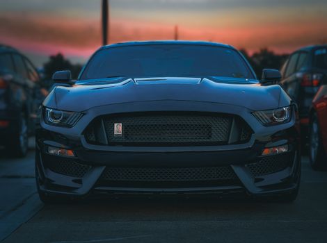 Front view of a sleek black sports car parked in a lot during a sunset, capturing its luxury and elegance.
