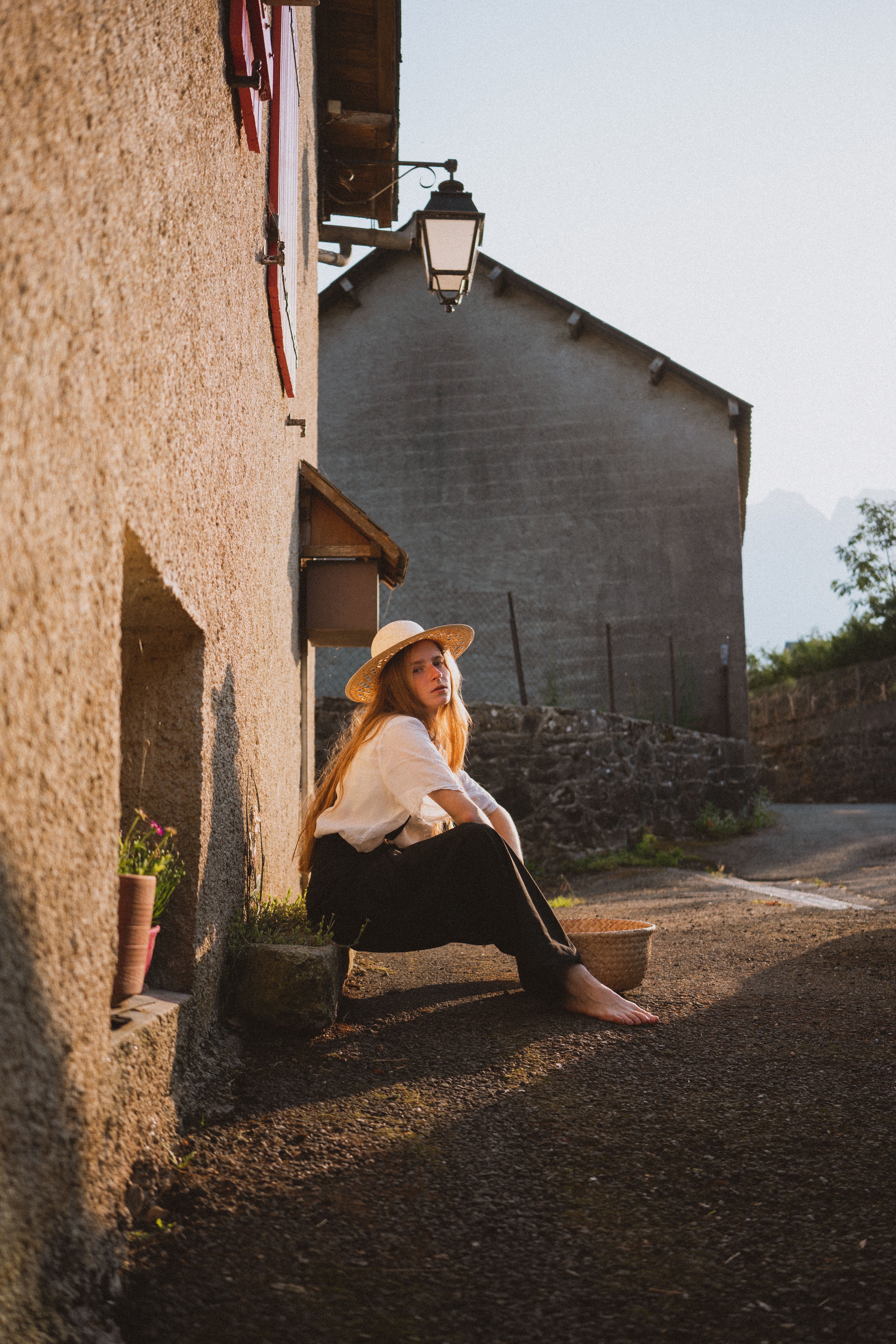 A woman in a straw hat sits outdoors, bathed in summer sunlight in a quaint village setting.
