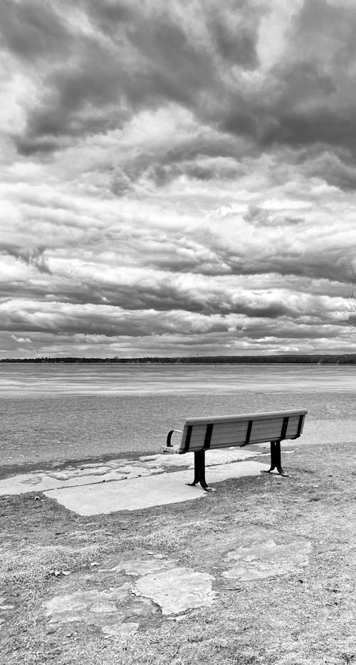 Grayscale Photo of a Bench on Beach under the Cloudy Sky