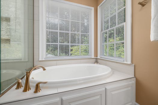 Bright bathroom featuring a white bathtub, brass faucets, and large windows with blinds.