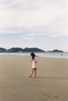 A young girl in a pink dress walking on a sandy beach with distant hills.