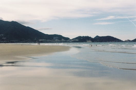 Peaceful beach scene with mountains and shallow waves under a blue sky.