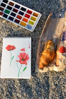 Creative flat lay featuring watercolor paints, a painted flower sketch, and a croissant.
