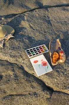 Watercolors, croissant, and sun hat on sandy beach rock, perfect for a creative outdoor escape.