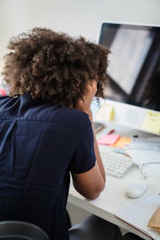 An adult woman with curly hair experiencing stress at her office desk, focusing on computer.