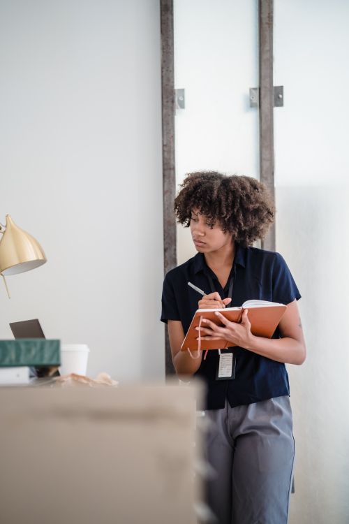 Free Young businesswoman with curly hair standing in office, writing notes in notebook, illustrating a professional and focused work environment. Stock Photo