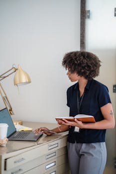 African American businesswoman standing at desk using laptop and holding a notebook in a modern office.