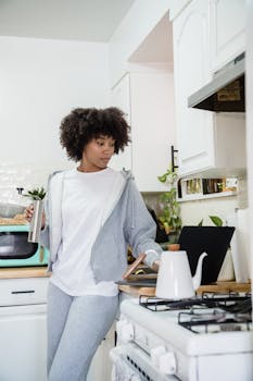Young African American woman in a cozy kitchen using a laptop, drinking coffee.