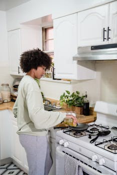 African American woman preparing food indoors, adding oil to pan in modern kitchen.