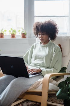 Focused African American woman using a laptop while relaxing indoors in a cozy armchair.