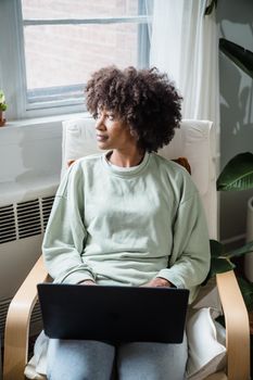 African American woman sitting in armchair using laptop, looking thoughtfully out the window.