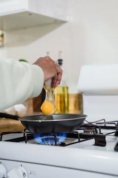 Close-up of hand cracking an egg into a frying pan on a lit stove.