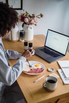 A woman enjoys breakfast with coffee and eggs while working on her laptop at home.