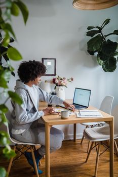A woman works on her laptop at a home office table surrounded by plants and natural light.