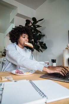 African American woman eating breakfast while using a laptop at the table indoors.
