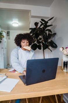 Young woman working on a laptop while eating breakfast at home, showcasing multitasking.