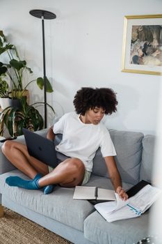 A young woman multitasks with a laptop and documents on a sofa in a cozy living room setting.