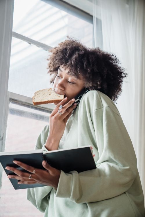 Free African American woman multitasking by eating toast, talking on phone, and reading. Stock Photo