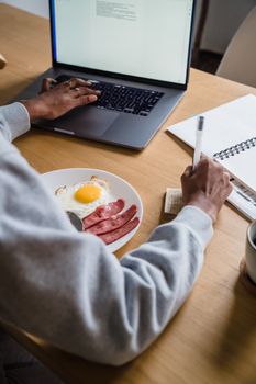 A person writes while having breakfast and using a laptop, showcasing a modern lifestyle.