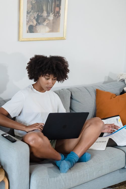 Free Woman enjoying remote work setup at home, sitting on a couch with her laptop and books. Stock Photo