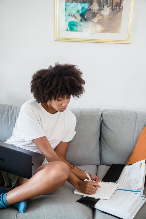 Free Woman sitting on a sofa, multitasking with a laptop and notepad while working from home. Stock Photo