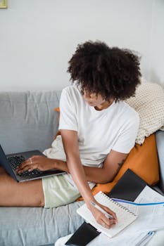 Woman working remotely on a laptop at home while taking notes, showcasing modern learning style.
