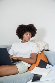 African American woman working on a laptop while sitting on a sofa in a relaxed home environment.