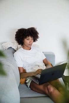 A focused woman sits on a sofa with a laptop, working remotely in a comfortable home setting.