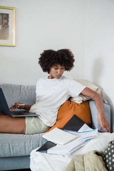A woman working remotely on a laptop from a comfortable sofa at home with documents scattered around.