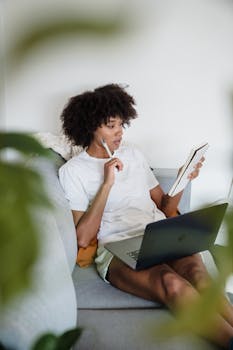 Focused young woman studying at home with a laptop and notepad, sitting comfortably on a couch.