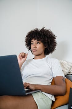A young woman with curly hair using a laptop while pondering a thought in a cozy home setting.