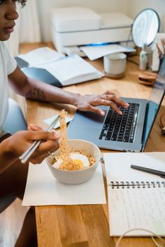 Young woman eats noodles while working remotely on a laptop at home.