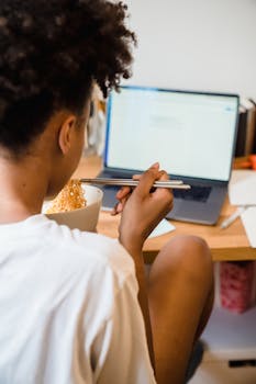 Woman eating noodles with chopsticks while using a laptop at a home desk.